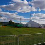 Temporary white tents and equipment on a grassy field with traffic cones and metal barriers
